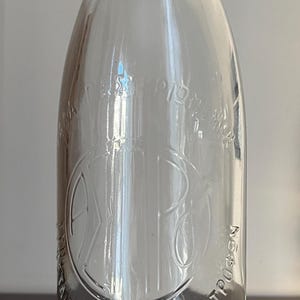 Puede incluir: Una botella de leche de vidrio transparente con el texto "ΑΓΡΟΤΙΚΟΣ ΑΤΕ ΚΑΟΤΡΥΓΩΝ ΑΣΠΡΟΥ ΓΑΛΑΚΤΟΣ" en relieve en el lateral.