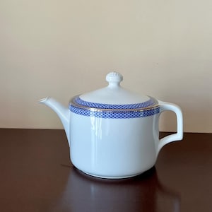 May include: White ceramic teapot with a lid and handle. The lid and rim feature a blue and gold patterned design. The teapot has a spout for pouring and is set on a dark brown surface.