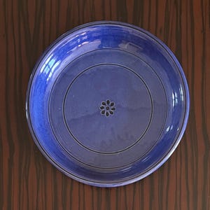Op de afbeelding: Een ronde, blauwe keramische plaat met een donkerblauwe rand en een centraal bloemmotief. De plaat heeft concentrische cirkels en een glanzende afwerking, geschikt voor het serveren van voedsel. De plaat staat tegen een donkere houtnerf achtergrond.