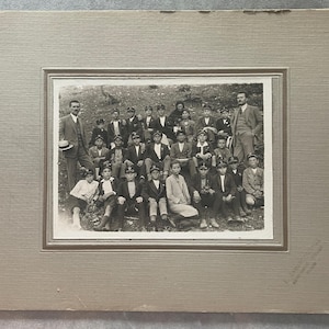 May include: A vintage black and white photograph featuring a group of people, likely a school or club, posing outdoors. The subjects are dressed in formal attire, including suits and caps. The photograph is mounted on a textured, light-colored mat.