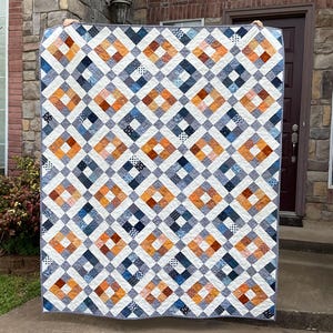 Op de afbeelding: Een quilt met een ruitpatroon in tinten blauw, oranje en wit. De quilt is gemaakt van vierkanten van verschillende stoffen en is gestikt met wit garen.