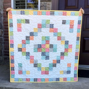 May include: A white quilt with a colorful geometric pattern. The quilt has a yellow border with a repeating pattern of circles.