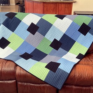 May include: A blue, green, black, and white patchwork quilt with a geometric pattern. The quilt is folded and draped over a brown leather couch.