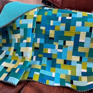 Może przedstawiać: Kołdra w patchworku w kolorze niebieskim i zielonym z czarno-białym wykończeniem. Kołdra jest złożona i leży na brązowej skórzanej kanapie.