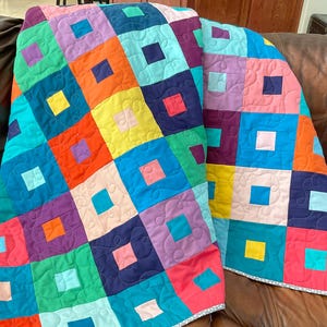 May include: A colorful quilt with a patchwork design. The quilt features squares in various colors, including blue, green, orange, pink, and purple. Each square has a smaller square in a contrasting color in the center. The quilt is draped over a brown leather couch.