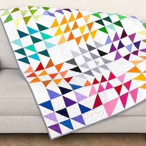May include: A quilt featuring a geometric star pattern composed of numerous small triangles in a vibrant rainbow spectrum of colors including red, orange, yellow, green, blue, and purple, all set against a white background with gray trim.