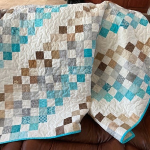 以下が含まれることがあります： 白い背景に青、茶色、グレーの布の正方形が並んだパッチワークキルト。キルトは半分に折り畳まれ、茶色の革のソファの上に置かれています。