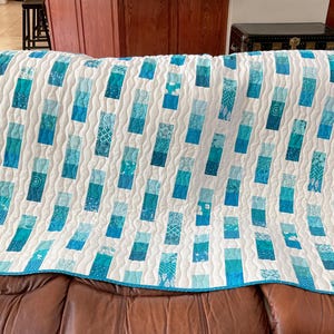 May include: A handmade quilt featuring a white backdrop with vertical columns of blue and teal patterned fabric rectangles. The quilt has a teal border and is draped over a brown leather sofa.