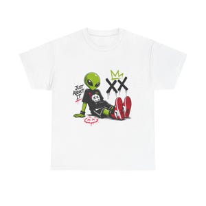 May include: White t-shirt featuring a cartoon alien in a black outfit and red sneakers. The alien is sitting with the text "JUST ABDUCT IT" and graffiti-style graphics. A green crown and "XX" are also visible.