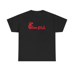 Op de afbeelding: Zwart T-shirt met een rode "Alien-fil-A" afbeelding. Het ontwerp vervangt het gebruikelijke logo door een alienhoofd. Het shirt is gemaakt van een zacht materiaal en heeft een klassieke ronde hals.