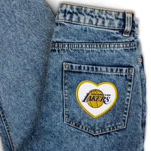 May include: Acid-wash blue denim jeans featuring a high-waisted design with belt loops and a single back pocket. A heart-shaped patch is stitched onto the pocket, displaying a yellow basketball and the text Los Angeles Lakers in purple.