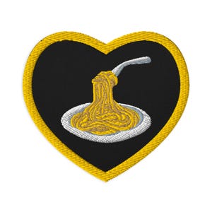 May include: A heart-shaped embroidered patch with a black background and a thick yellow border. The center features a white plate holding a mound of yellow spaghetti with a silver fork lifting a portion of the pasta strands upward.