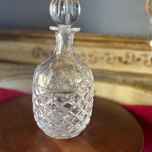 May include: A clear glass decanter with a diamond-cut design and a round stopper. The decanter is on a wooden surface. The background features a gold decorative element and a red textile.