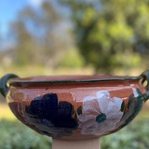 May include: A hand-painted terracotta bowl with a wide rim and two green handles. The bowl features a floral design with white and navy blue flowers. The rim is painted green, and the bowl is set on a beige pedestal.