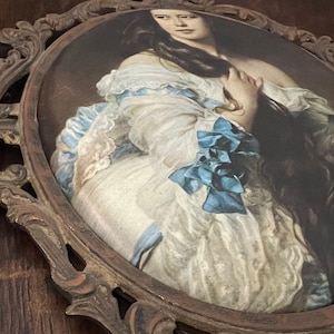 Puede incluir: Un marco ovalado adornado con un dise&ntilde;o detallado e intrincado. El marco est&aacute; hecho de metal oscuro y presenta una superficie texturizada en relieve. Dentro del marco hay un retrato vintage de una mujer con cabello largo y oscuro que lleva un vestido blanco con una faja azul.