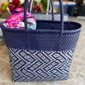 May include: A woven tote bag featuring a dark navy blue upper section and a lower section with a geometric pattern in navy blue and silver. The bag has two matching navy blue braided handles and contains a cluster of bright pink artificial flowers.