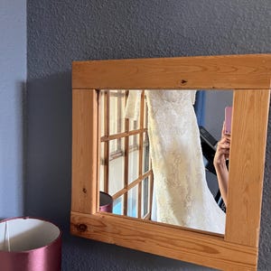 May include: A wooden-framed mirror reflecting a white wedding dress with lace detailing. The mirror is mounted on a dark gray wall, and a burgundy lamp is visible in the foreground. The reflection also shows a window.