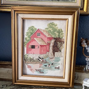May include: A framed cross-stitch embroidery depicting a red water mill with a wooden wheel situated beside a pond. Several ducks swim in the water in the foreground. The artwork is encased in a gold-toned frame with a cream-colored inner border.