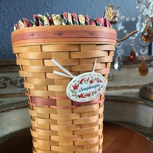 May include: A woven basket with a light brown hue, featuring a decorative rim and a pink band. The basket has a tag that reads "Snapdragon" with floral accents. The top edge is lined with colorful fabric. The basket is on a wooden surface.