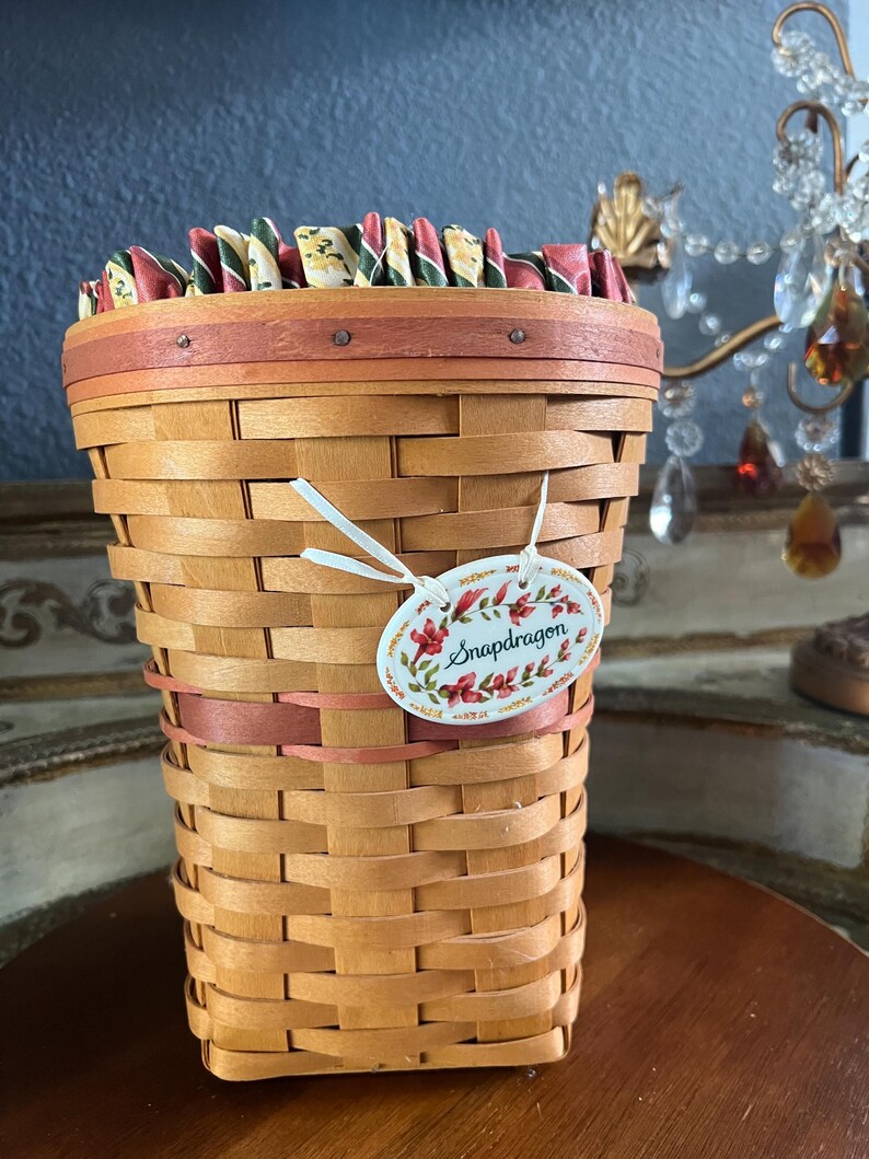 May include: A woven basket with a light brown hue, featuring a decorative rim and a pink band. The basket has a tag that reads "Snapdragon" with floral accents. The top edge is lined with colorful fabric. The basket is on a wooden surface.