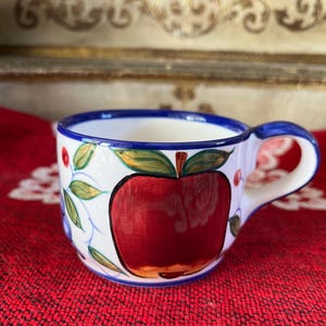 May include: A small, white ceramic teacup with a large red apple design. The cup has a blue rim and handle, with green leaves and red berries painted around the apple. The cup is sitting on a red surface.