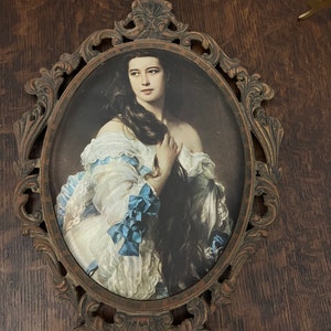 Puede incluir: Un retrato ovalado de una mujer con el pelo largo y oscuro que lleva un vestido blanco con cintas azules, enmarcado en un marco de metal dorado adornado.