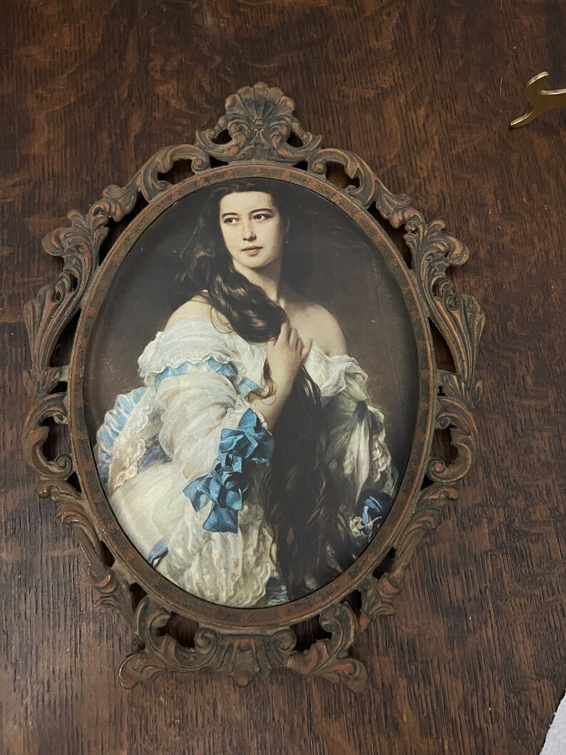 Puede incluir: Un retrato ovalado de una mujer con el pelo largo y oscuro que lleva un vestido blanco con cintas azules, enmarcado en un marco de metal dorado adornado.