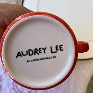 K&ouml;nnte beinhalten: Eine rot umrandete Kaffeetasse mit dem Text "AUDREY LEE" in Schwarz und "for ANTHROPOLOGIE" darunter, vor einem wei&szlig;en Innenraum. Im Hintergrund befindet sich ein wei&szlig;er Teller.
