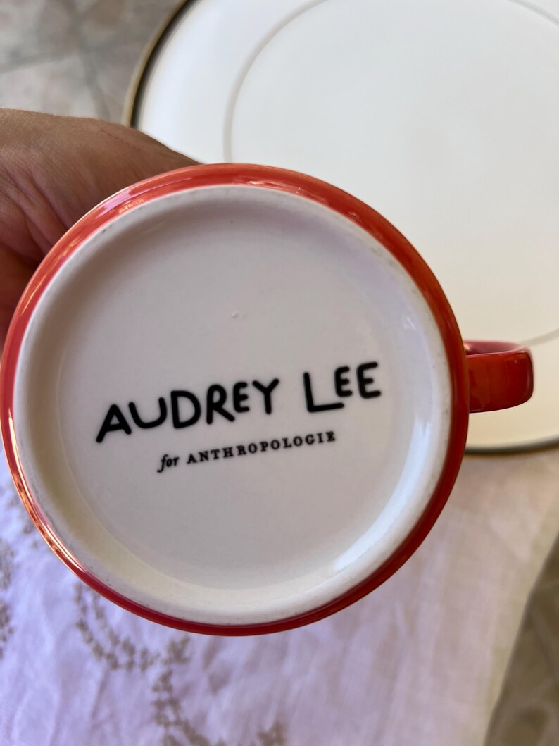 K&ouml;nnte beinhalten: Eine rot umrandete Kaffeetasse mit dem Text "AUDREY LEE" in Schwarz und "for ANTHROPOLOGIE" darunter, vor einem wei&szlig;en Innenraum. Im Hintergrund befindet sich ein wei&szlig;er Teller.