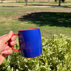 K&ouml;nnte beinhalten: Eine blaue Keramiktasse mit rotem Innenraum und Henkel. Die Tasse hat eine strukturierte Oberfl&auml;che und wird vor einem gr&uuml;n-wei&szlig; belaubten Busch gehalten. Der Hintergrund zeigt eine grasbewachsene Fl&auml;che mit B&auml;umen.