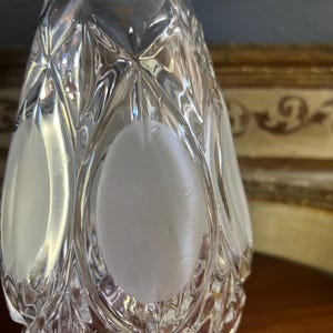 May include: A clear, faceted glass bottle with a diamond pattern and frosted oval accents. The bottle has a teardrop shape and is likely a decorative item or a perfume bottle.