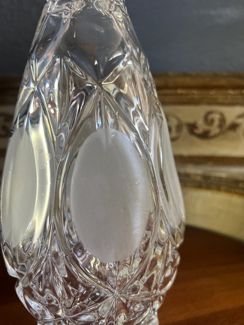 May include: A clear, faceted glass bottle with a diamond pattern and frosted oval accents. The bottle has a teardrop shape and is likely a decorative item or a perfume bottle.