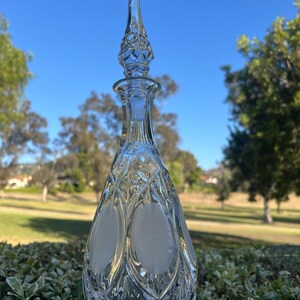 May include: A clear glass decanter with a diamond pattern and a pointed stopper. The decanter has two oval frosted sections on the front. The background shows a blue sky and green foliage.