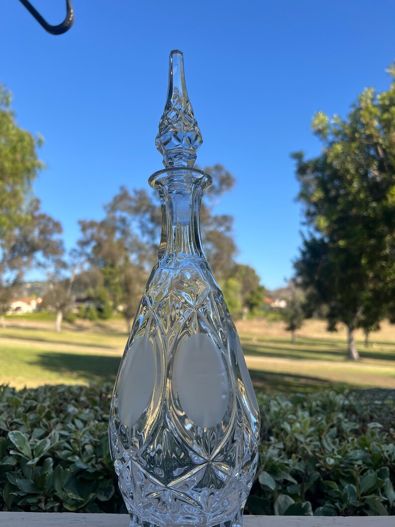 May include: A clear glass decanter with a diamond pattern and a pointed stopper. The decanter has two oval frosted sections on the front. The background shows a blue sky and green foliage.
