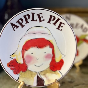 May include: A white ceramic plate with a brown rim and the words "APPLE PIE" in dark red. The plate features a cartoon illustration of a girl with red hair and a yellow hat. The plate is displayed on a gold-colored stand.