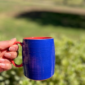 K&ouml;nnte beinhalten: Eine blaue Keramiktasse mit rotem Innenraum und Henkel. Die Tasse hat ein strukturiertes, geripptes Design auf der Au&szlig;enseite. Der Henkel ist rot, und die Tasse wird vor einem verschwommenen gr&uuml;nen Hintergrund gehalten.