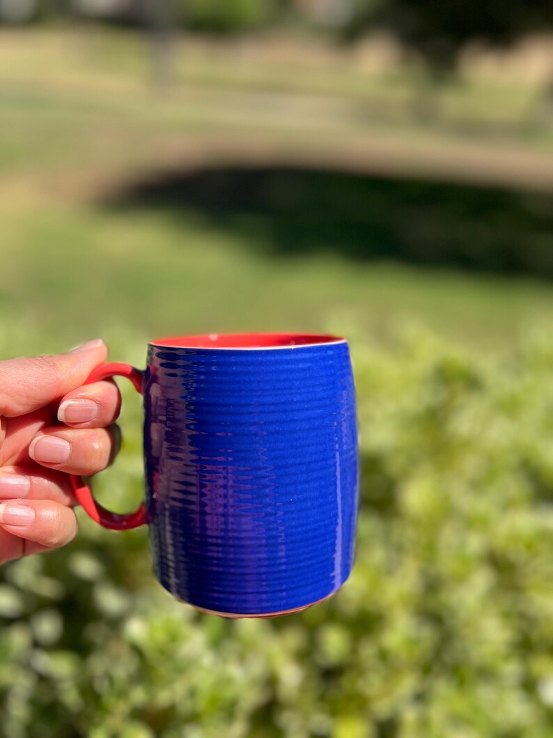 K&ouml;nnte beinhalten: Eine blaue Keramiktasse mit rotem Innenraum und Henkel. Die Tasse hat ein strukturiertes, geripptes Design auf der Au&szlig;enseite. Der Henkel ist rot, und die Tasse wird vor einem verschwommenen gr&uuml;nen Hintergrund gehalten.