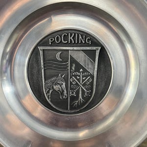 May include: A silver pewter plate with a raised design of a coat of arms. The coat of arms features a horse's head, a crescent moon, and a tree with a cross. The plate is inscribed with the word "POCKING".