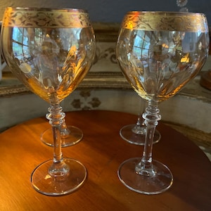 May include: Four clear wine glasses with amber-colored bowls and ornate gold rims. The stems are clear with decorative detailing. The glasses are arranged on a round, wooden table.