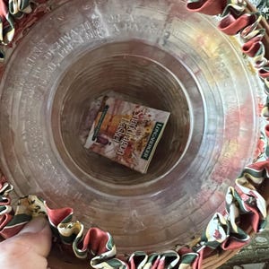 May include: A round, clear glass basket with a decorative rim of red, green, and cream-colored fabric. The basket contains a small book titled "Shadow Dragon Basket" by Longaberger. The basket is made of woven wood.