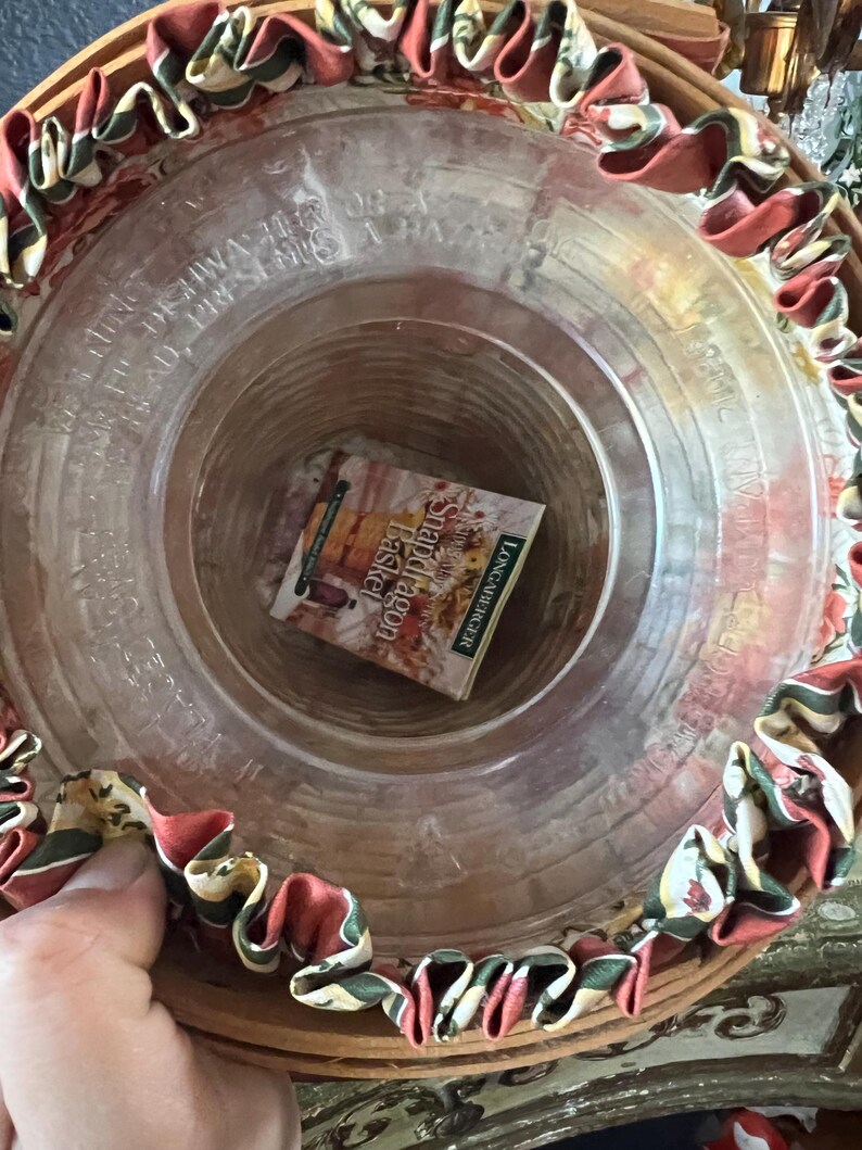 May include: A round, clear glass basket with a decorative rim of red, green, and cream-colored fabric. The basket contains a small book titled "Shadow Dragon Basket" by Longaberger. The basket is made of woven wood.
