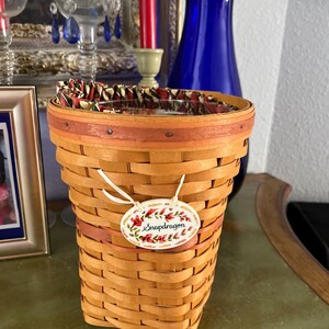 May include: A woven basket with a floral tag that reads "Snapdragon." The basket is light brown with a dark brown rim and a patterned fabric lining the top. The tag has a floral design with red and green accents.
