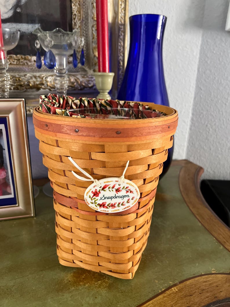 May include: A woven basket with a floral tag that reads "Snapdragon." The basket is light brown with a dark brown rim and a patterned fabric lining the top. The tag has a floral design with red and green accents.