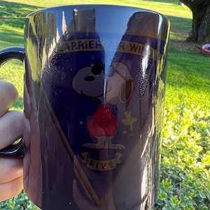 May include: A navy blue ceramic mug featuring Snoopy from Peanuts, with the text "CARRIER AIR WING FIVE". The mug has a black handle and a design that includes a red emblem and a cartoon airplane.