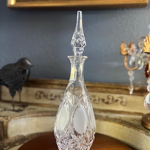 May include: A clear glass perfume bottle with a diamond-cut design and a long, pointed stopper. The bottle has a teardrop shape with frosted oval accents. The bottle is displayed on a brown surface.