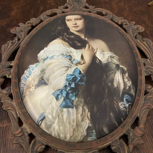 Puede incluir: Un retrato enmarcado de una mujer con el pelo largo y oscuro que lleva un vestido blanco con detalles azules. El marco es ornamentado y de metal.