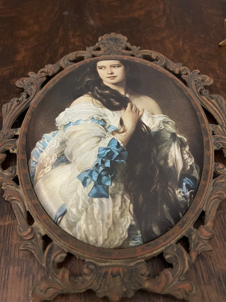Puede incluir: Un retrato enmarcado de una mujer con el pelo largo y oscuro que lleva un vestido blanco con detalles azules. El marco es ornamentado y de metal.