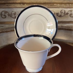 May include: A white teacup and saucer with a black and silver trim. The cup has a rounded shape and a handle. The saucer is flat with a raised edge.