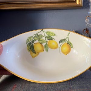 May include: Oval serving dish with a white interior and a yellow rim. The dish is decorated with a lemon branch design featuring yellow lemons and green leaves. The dish is likely made of ceramic.