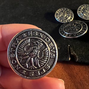Może przedstawiać: Srebrny metalowy guzik z wytłoczonym wzorem orła i otaczającym tekstem. Guzik jest trzymany w dłoni, z kilkoma innymi guzikami w tle. Guziki mają szczegółowy, antyczny wygląd.